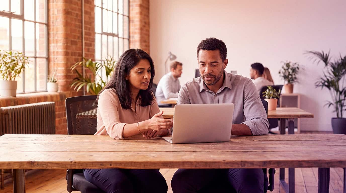 Two colleagues at a desk in a modern open-plan office, reviewing content on a laptop