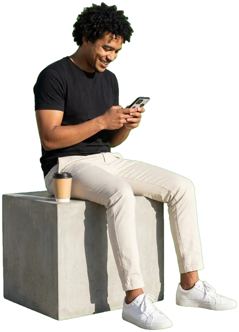 A young man sitting on a concrete block in the sun, grinning at his phone, a takeaway coffee beside him